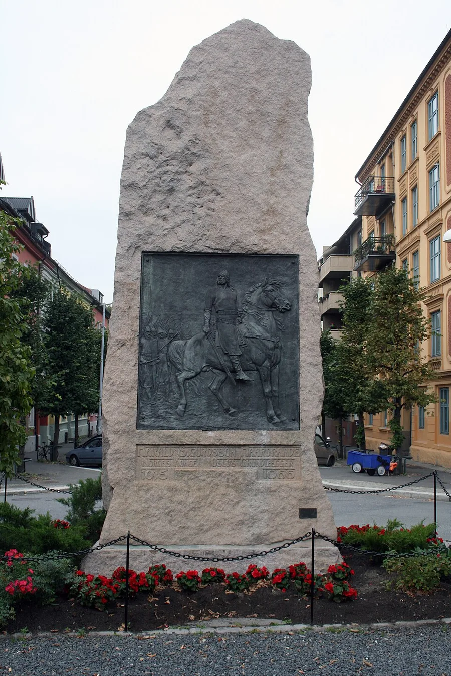 A tall rough-hewn granite bauta on Harald Hårdrådes plass, Oslo, with a bronze relief of a helmeted, caped man leading a horse by the reins; the plaque below reads ‘Harald Sigurdsson Hardraade, Norges Konge, Oslos Grundlegger, 1015-1066,’ red flowers at its base.