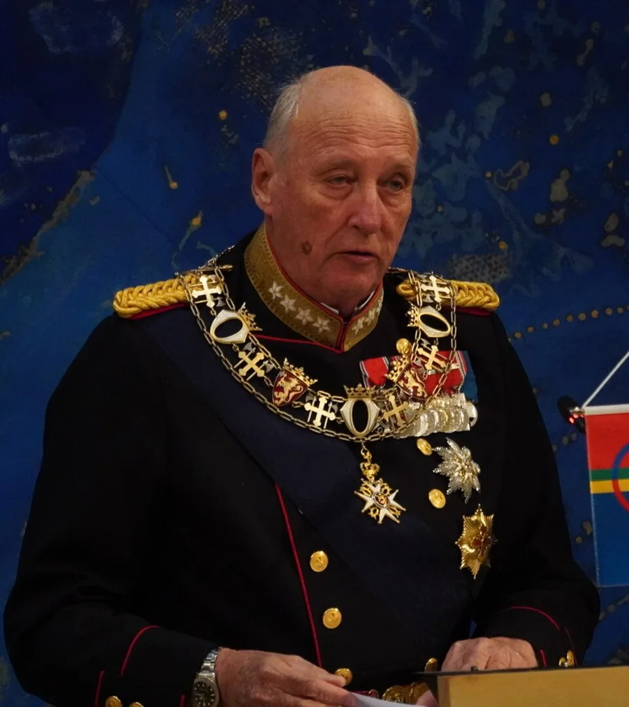 King Harald V of Norway in ceremonial dress uniform wearing the chain and star insignia of Norwegian royal orders, reading from notes at a lectern, against a blue mural backdrop with a Sámi flag visible over his shoulder.