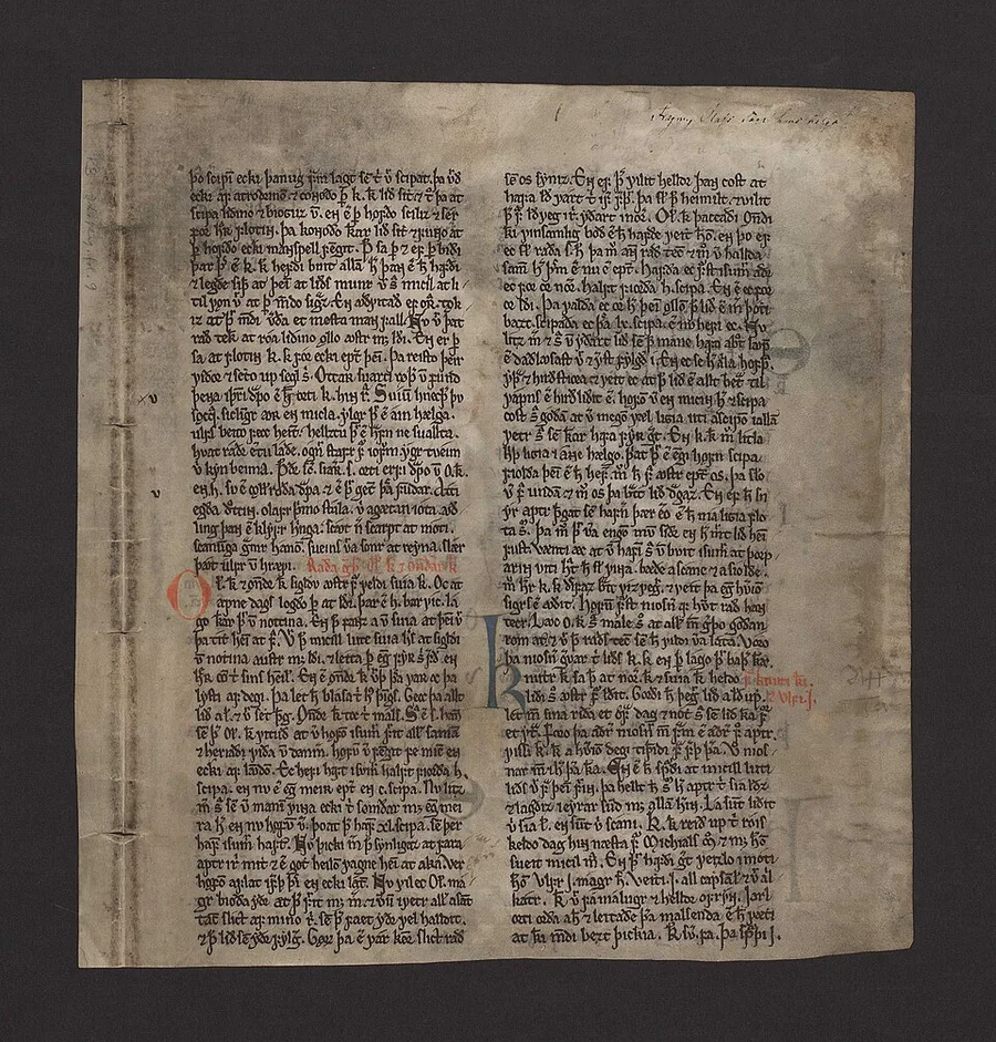 A single vellum leaf photographed on black: two dense columns of medieval Icelandic script in brown ink with red rubrics, a large red initial O and a blue initial K, and stitched repairs along the left edge — the Kringla leaf, sole survivor of the lost Kringla manuscript.