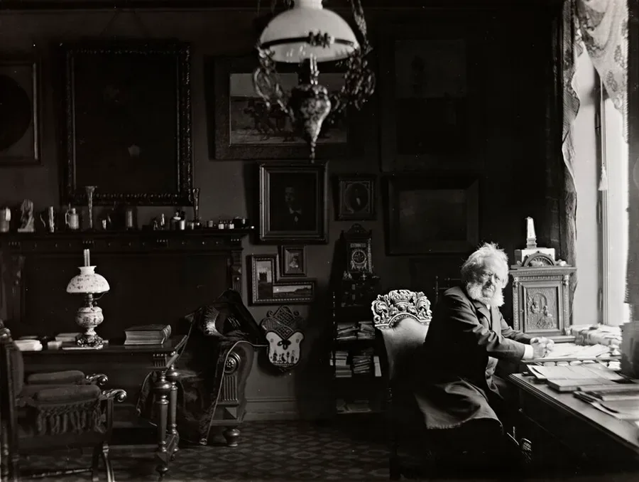 Photograph of an elderly, white-bearded Henrik Ibsen seated at a writing desk in a cluttered, dimly lit study lined with paintings, a hanging lamp, and a clock, glancing toward the camera.
