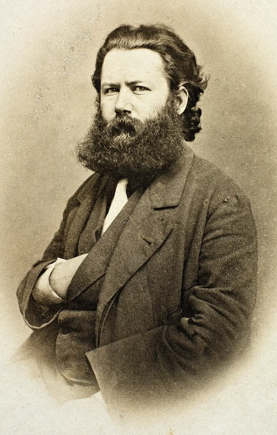 Sepia studio photograph of Henrik Ibsen in his mid-thirties, half-length with arms crossed, full dark beard and wavy swept-back hair, wearing a wide-lapelled coat.