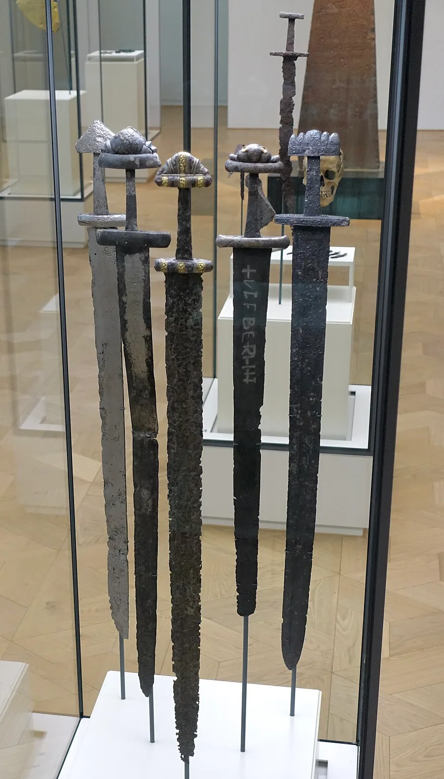 Five Viking-Age swords with ornamented hilts stand upright in a glass museum case, one blade inlaid ULFBERHT in lettering, with an out-of-focus human skull displayed in the case behind them.