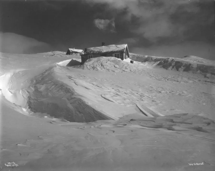 Black-and-white photograph of a turf-roofed timber cabin standing alone on a wind-carved snowdrift high above the treeline, a smaller outbuilding behind it and a dark, dramatic sky overhead.