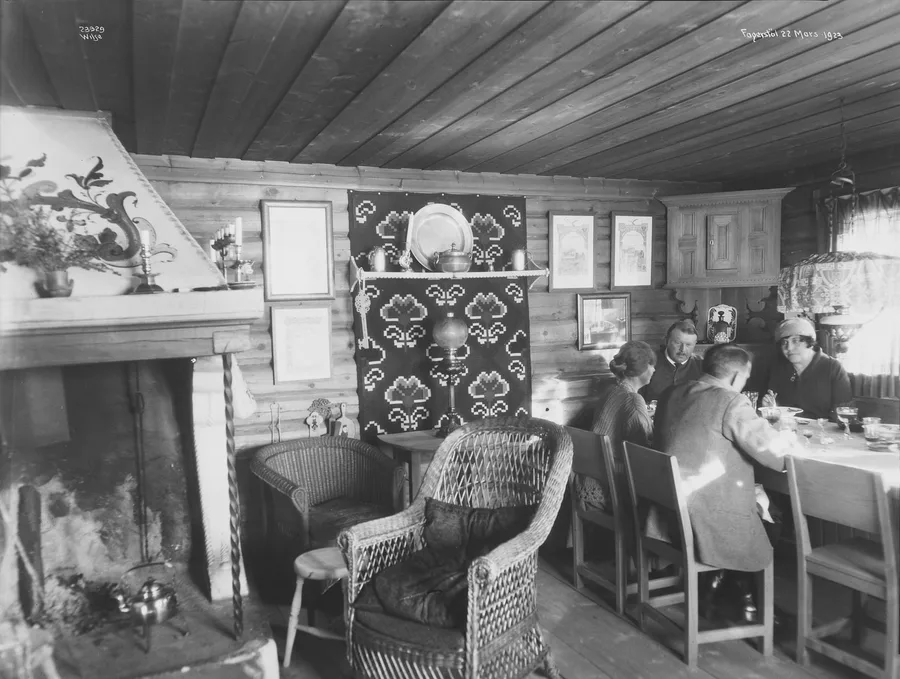 Black-and-white photograph of a log-walled cabin living room in 1923 — an open hearth with a rosemaling-painted hood and a kettle on the floor, wicker chairs, a patterned wall hanging, and four adults sharing a meal at a white-clothed table by the window.