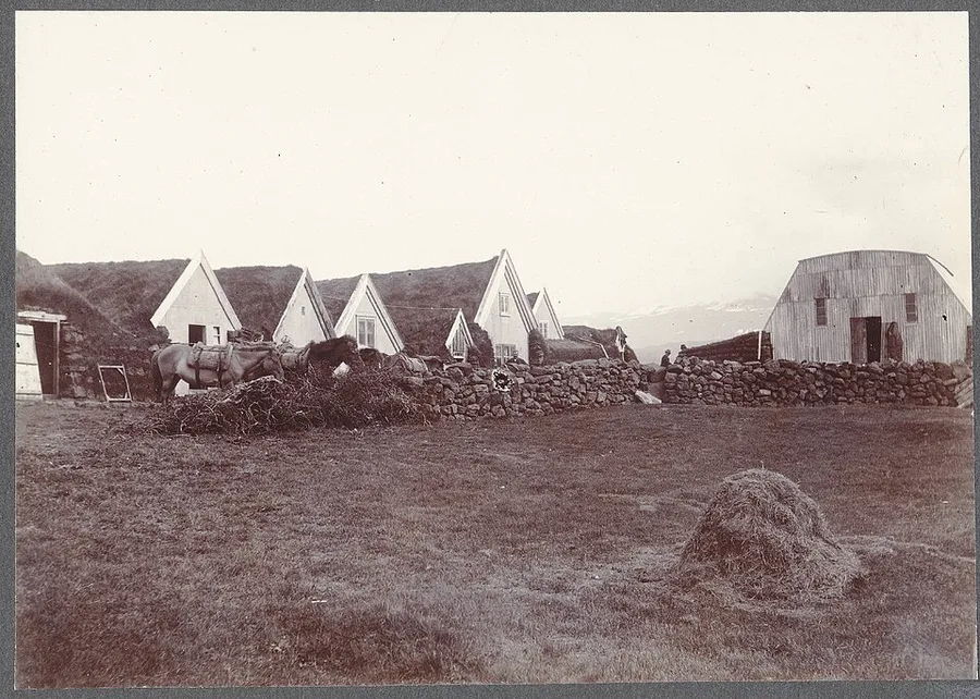 Sepia photograph from about 1900 of the farm at Galtalækur, Iceland: a row of white-gabled farmhouses built into turf and stone walls, pack ponies standing at the wall, a haycock in the home field, and the slopes of Hekla lost in haze behind.