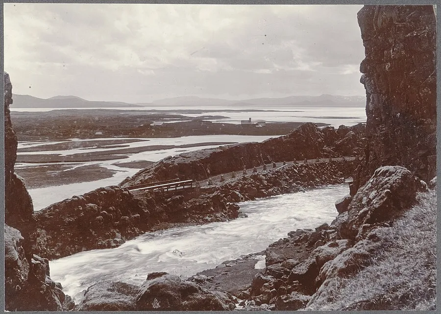 Sepia photograph from about 1900 looking out of the Almannagjá rift at Þingvellir: the Öxará river foaming past a small wooden footbridge in the gorge, with the assembly plain, the lake and distant fells beyond.