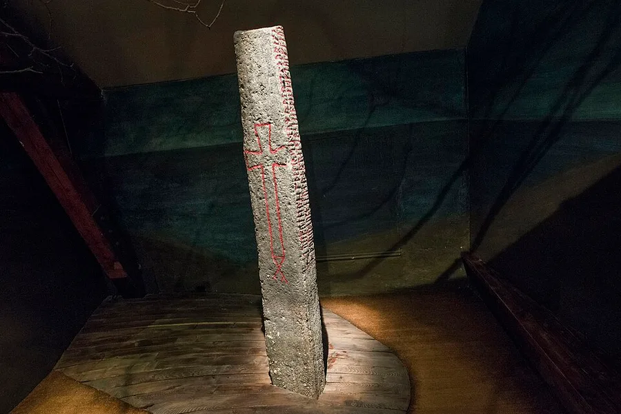 The Kuli Stone, a tall grey runestone carved with a Latin cross outlined in red and a vertical band of runic inscription, lit dramatically and displayed upright on a museum platform against a dark backdrop.