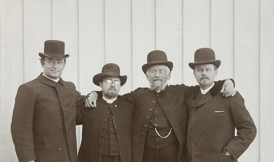 1904 black-and-white photograph of four clergymen in top hats and dark coats standing arm-in-arm against a white wooden wall, the two center men bearded and wearing clerical collars and a watch chain.