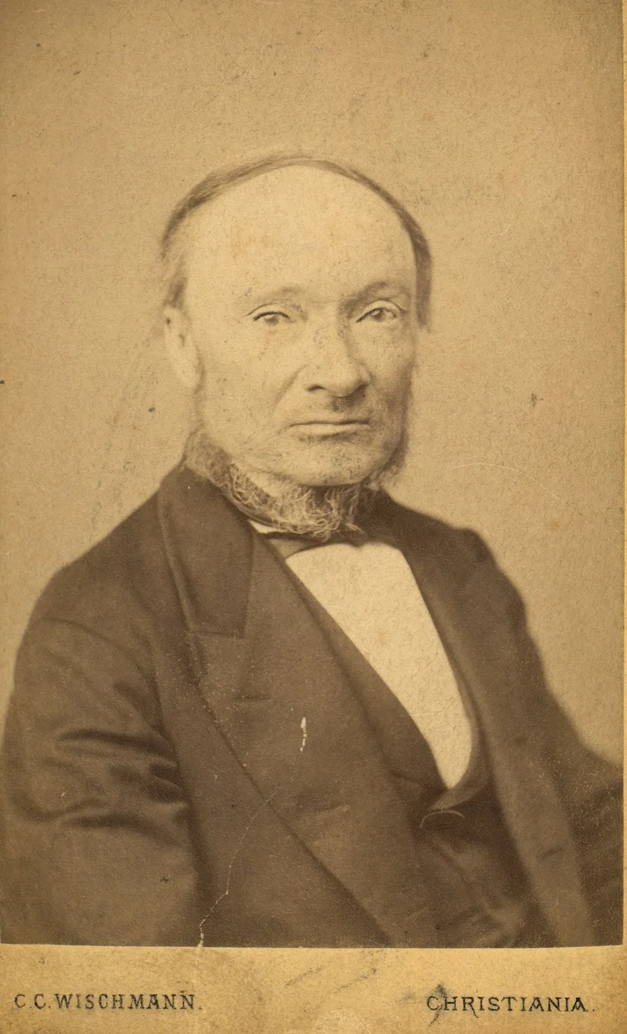 An 1871 sepia studio photograph of an older bearded man with receding hair, in a dark coat and bow tie, looking steadily toward the camera; the photographer's imprint "C.C. Wischmann, Christiania" below.