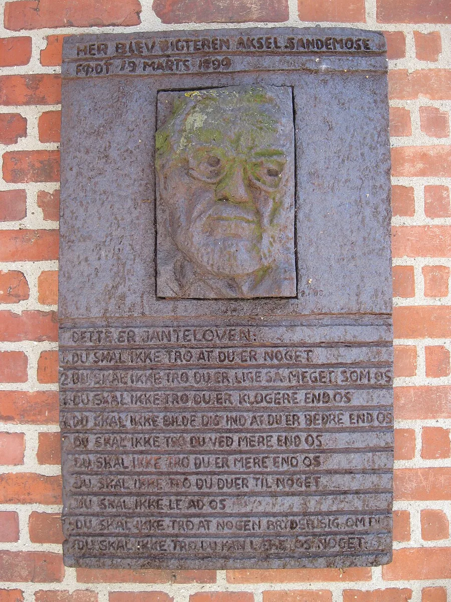 Weathered memorial plaque on a red-brick wall with a relief of Aksel Sandemose's bespectacled face; Danish text records that the writer was born here on 19 March 1899, and beneath the heading 'Dette er Janteloven' the ten rules are inscribed line by line.