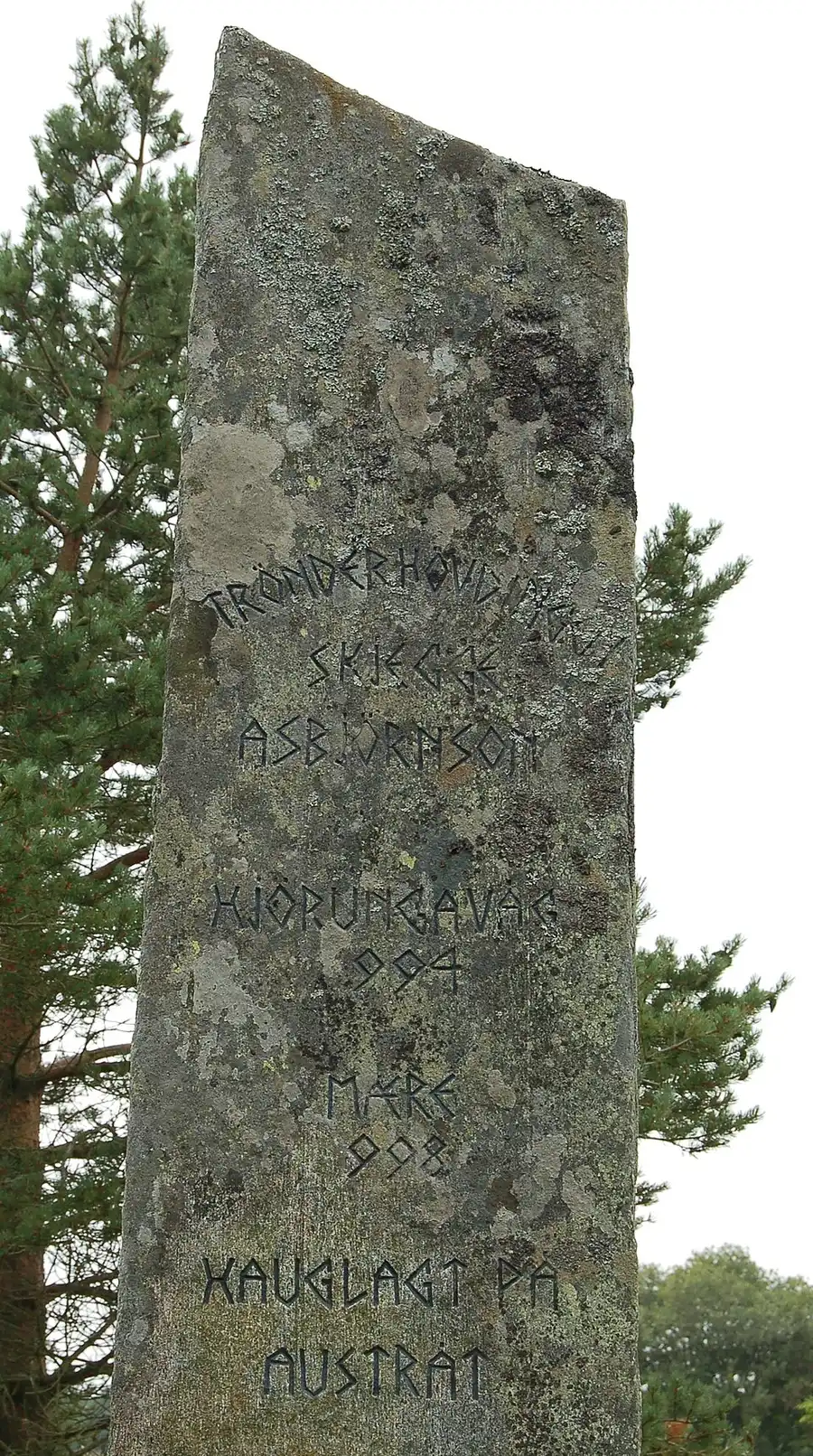 A tall, lichen-mottled memorial stone at Ørland, inscribed in runic-styled lettering to 'Trønderhøvdingen Skjegge Asbjørnson', naming Hjørungavåg and Mære and his mound-burial at Austrått ('hauglagt på Austråt'); pine boughs and a pale sky behind.