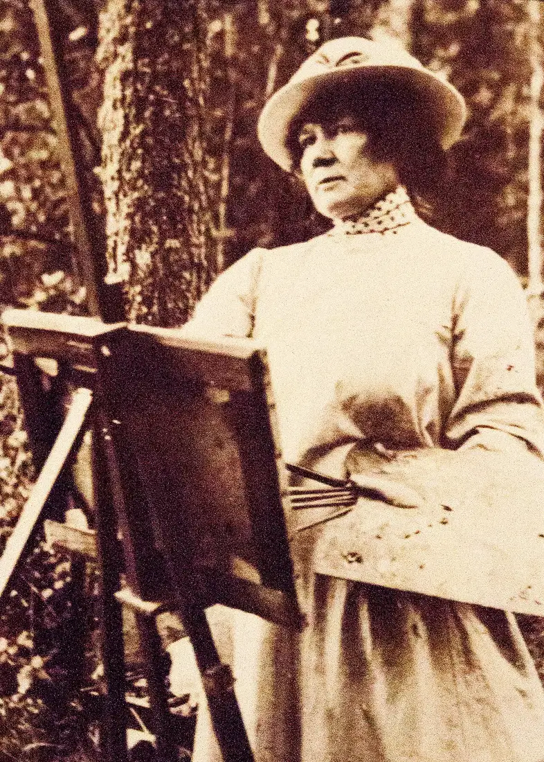 Sepia photograph of Jenny Nyström painting at an outdoor easel among trees — brimmed hat, high patterned collar, her palette and a fan of brushes on her arm.