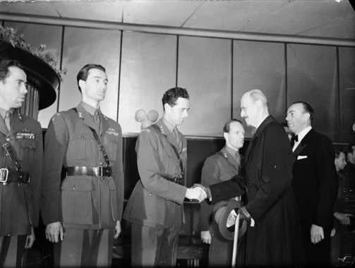 Black-and-white 1948 photograph of uniformed Gunnerside veterans lined up as King Haakon VII, in a dark overcoat and leaning on a cane, shakes hands with one of them outside a cinema.