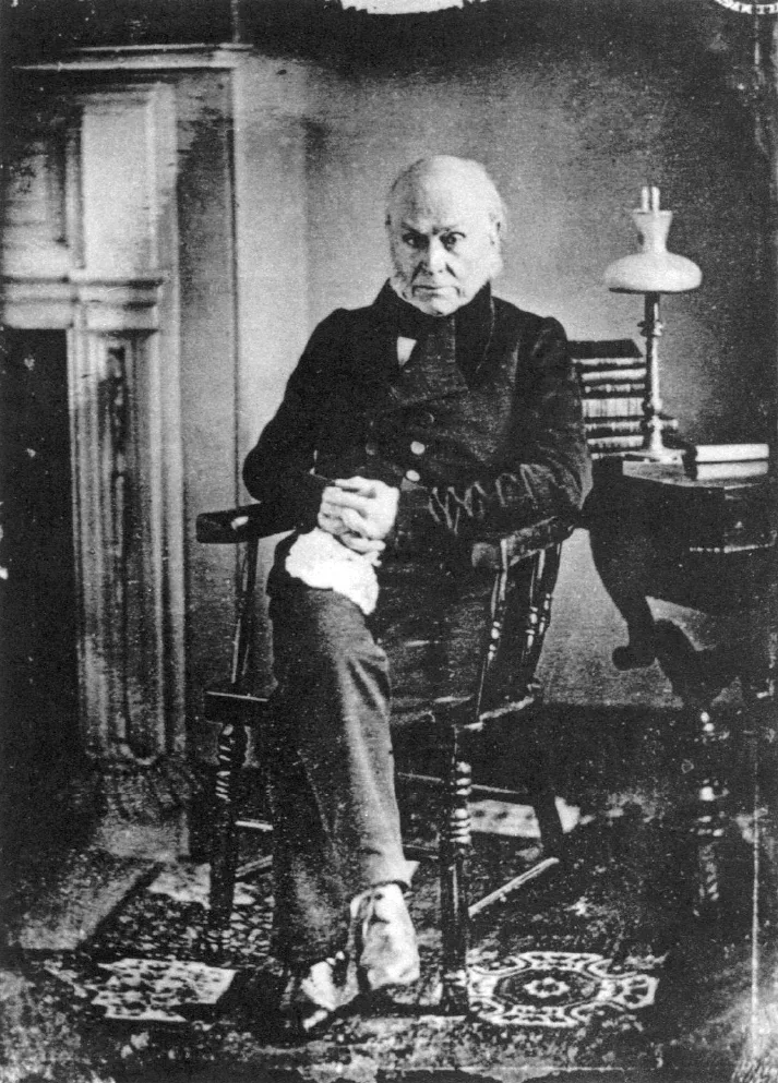 Full-length daguerreotype of an elderly, balding John Quincy Adams seated in a chair beside a fireplace, in a dark coat with a white collar, hands folded in his lap, a side table with books and an oil lamp beside him.