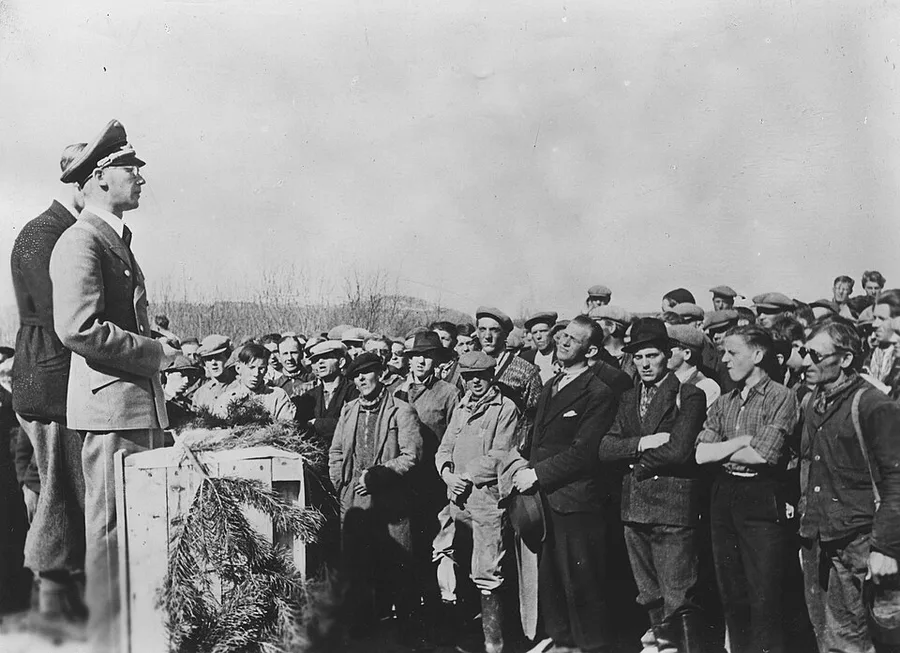 Black-and-white 1940 photograph of Josef Terboven in a military coat and cap, speaking from a fir-branch-decorated podium to a large crowd of Norwegian workingmen outdoors at Værnes garrison.