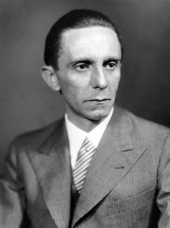 Black-and-white 1933 studio portrait of Joseph Goebbels, gaunt and dark-haired, in suit and striped tie, gazing slightly off-camera.