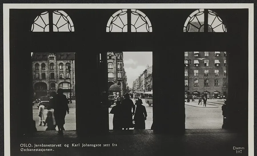 Black-and-white postcard seen through the three arched doorway bays of Oslo's old railway station: pedestrians and period buildings line Karl Johans gate toward Jernbanetorget, captioned 'Jernbanetorvet og Karl Johansgate sett fra Østbanestasjonen.'