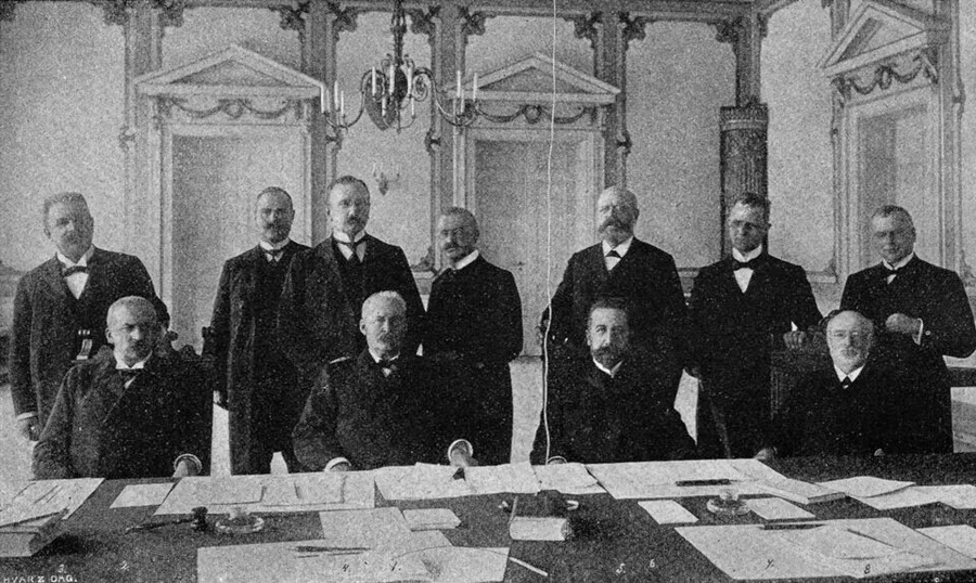Black-and-white group photograph of eleven mustached men in dark suits, some seated at a paper-covered table and others standing behind them in an ornate paneled hall — the Norwegian and Swedish delegations at the 1905 Karlstad negotiations.
