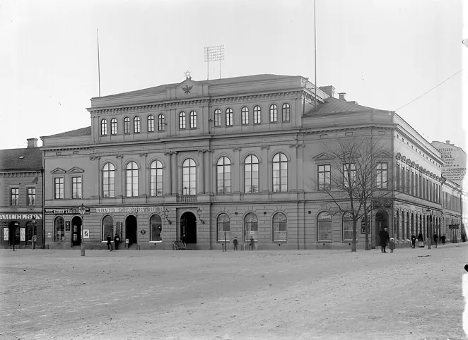 Black-and-white 1922 photograph of the Freemasons' lodge in Karlstad: a two-story neoclassical building with arched windows and a wide pediment facing the open market square, shopfronts along its ground floor and a few passers-by on the bare ground.