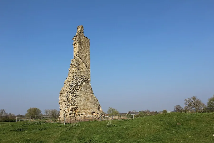 Color photograph of a single tall ruined stone wall — the surviving southeast corner of Kirkstead Abbey's church — standing alone in a green field under a clear blue sky.