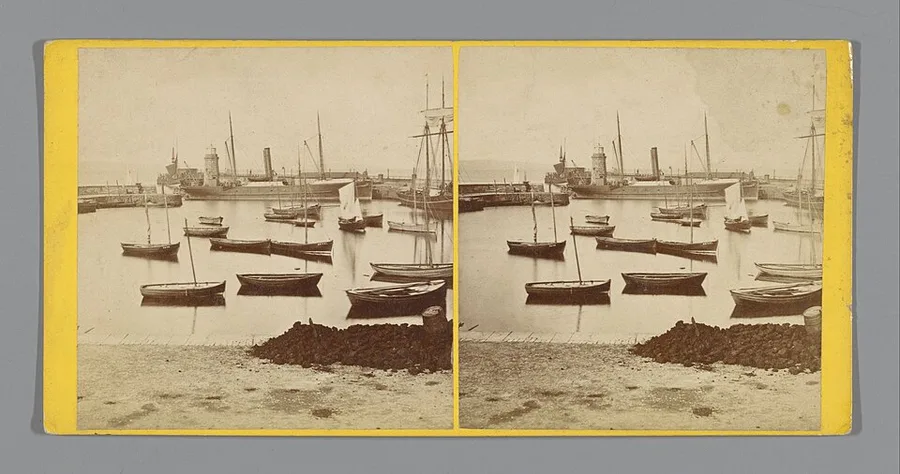 A sepia Victorian stereograph card showing twin identical views of Kirkwall harbour, small boats moored along the quay, a steamship and sailing vessels beyond, and the town's low stone buildings behind, mounted on a yellow card.