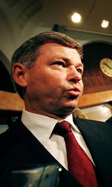 Color photograph, low angle, of a middle-aged man with combed gray-brown hair speaking, wearing a dark suit and red patterned tie, photographed indoors against a wood-beamed ceiling and a wall clock.