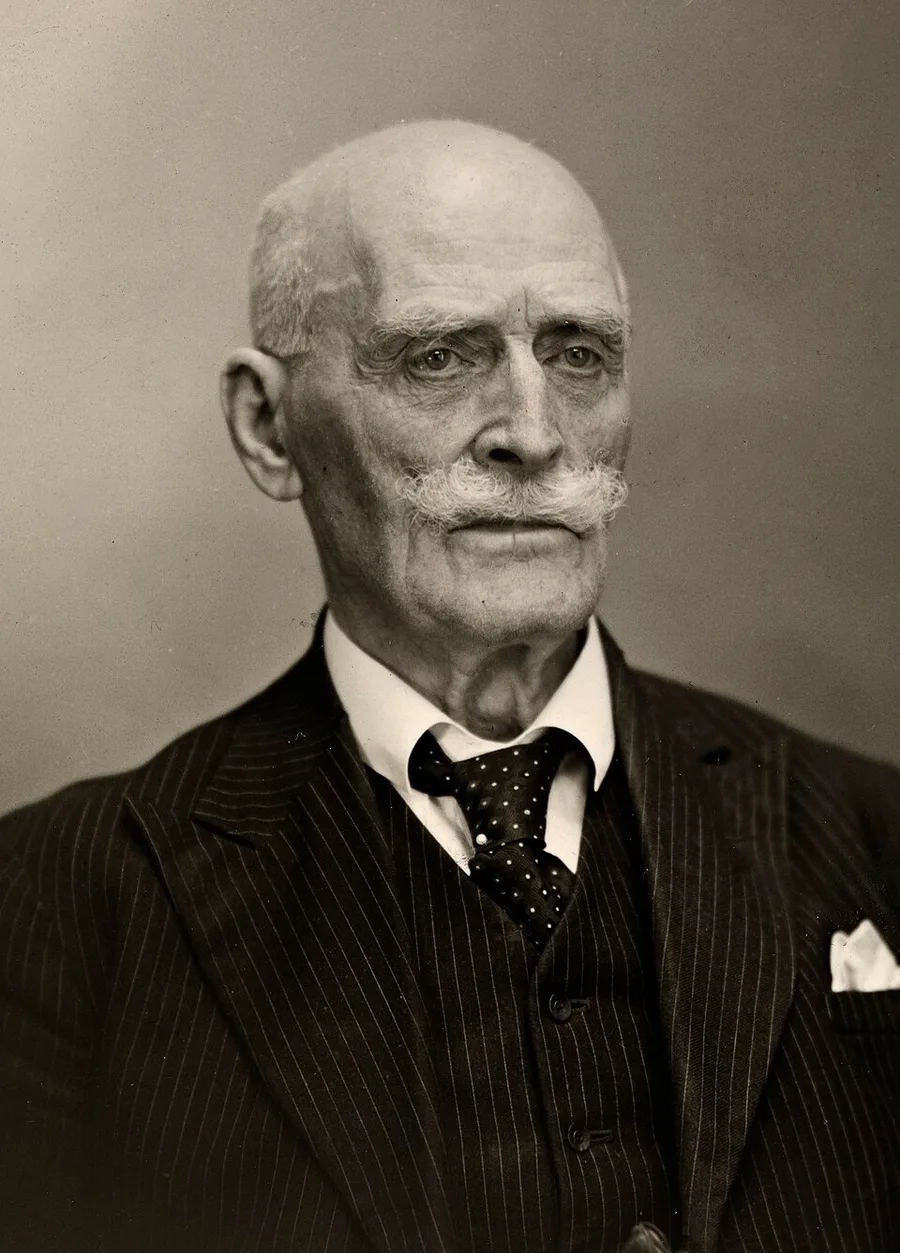 Black-and-white 1944 photograph of an elderly Knut Hamsun, bald with a heavy white mustache, in a pinstripe suit and polka-dot tie, facing the camera.