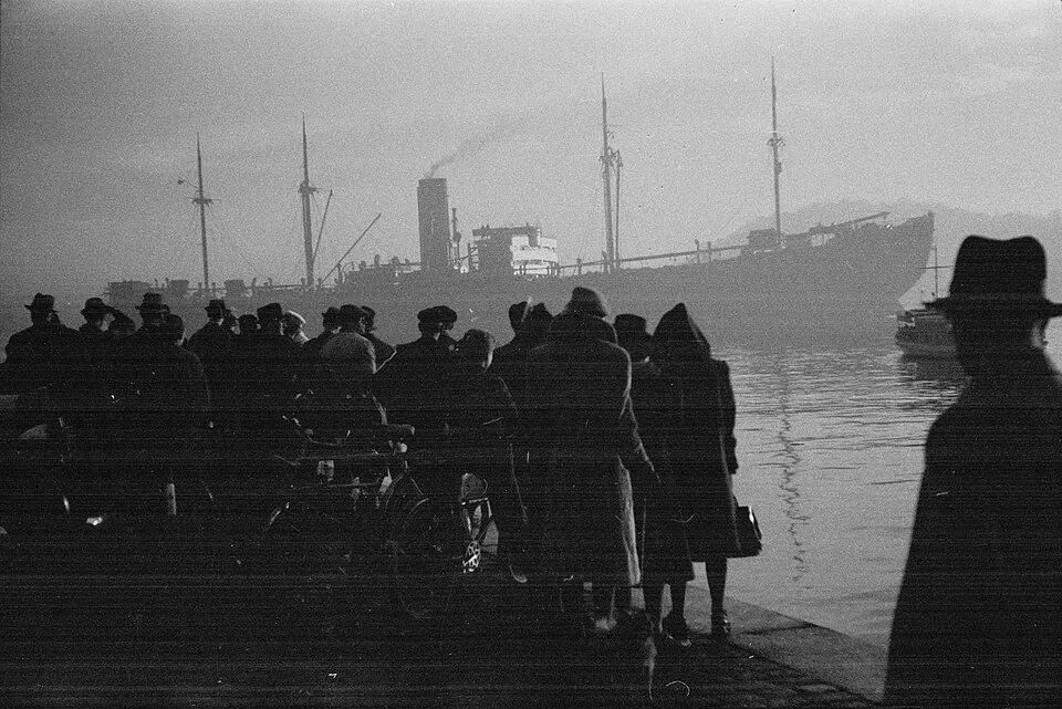 Black-and-white photograph, 26 November 1942: a crowd in hats and winter coats — Norwegian Jews on the Akershus quay in Oslo — stands with backs to the camera, a bicycle among them, as the dark cargo ship Donau lies in the harbor haze; a man in a fedora is silhouetted at right.