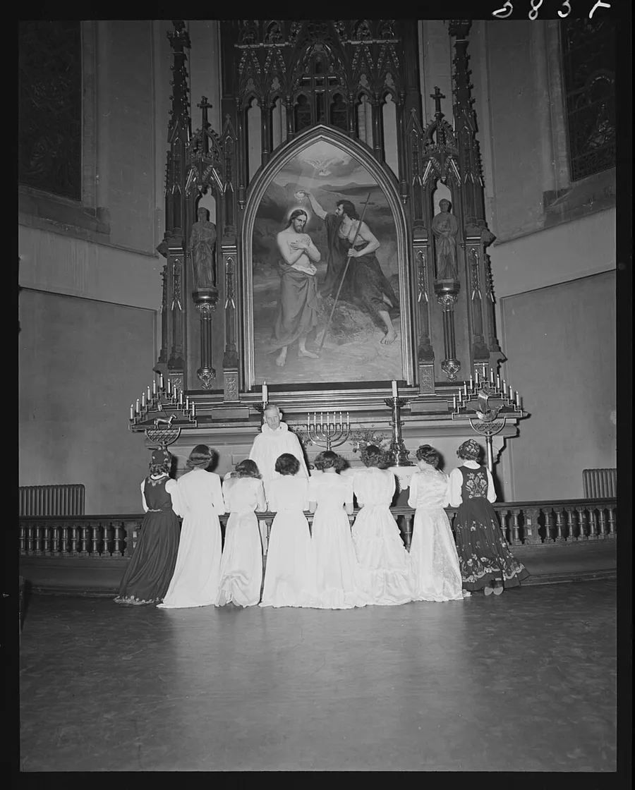 A 1948 black-and-white photo of confirmands in white dresses kneeling at the altar rail of Trefoldighetskirken, a priest standing before an ornate Gothic-revival altarpiece depicting Christ's baptism.