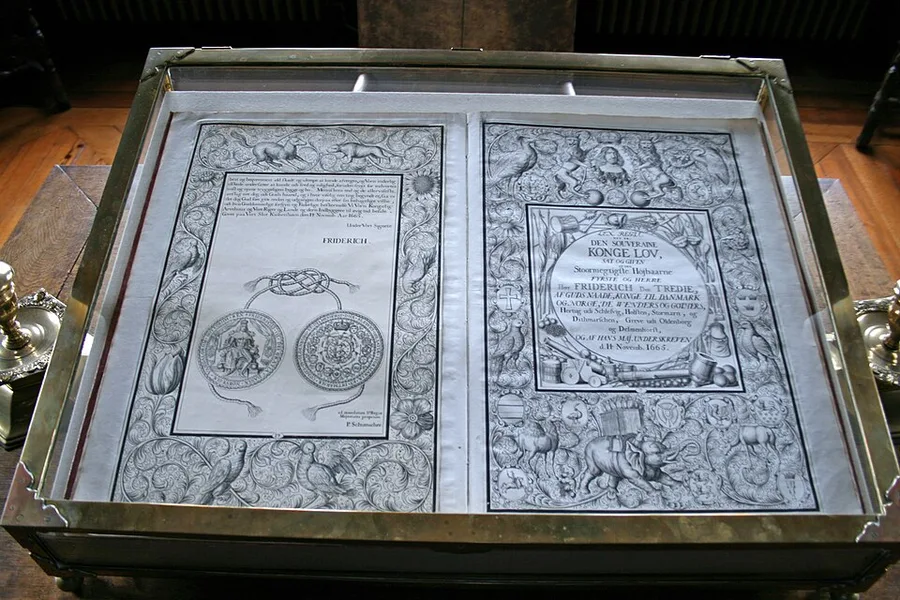 Photograph of the 1665 Kongeloven displayed open in a glass museum case at Frederiksborg Castle: two ornately bordered pages with engraved seal images on the left leaf and dense period lettering and royal titulature on the right.