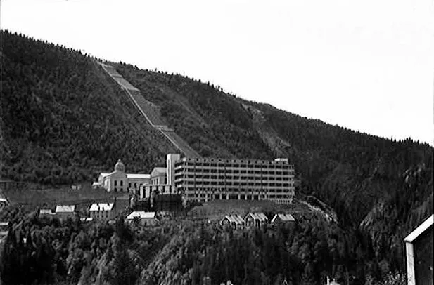 Black-and-white 1935 photograph of the Vemork hydroelectric plant near Rjukan: a long multi-story building with rows of arched windows built into a steep, forested valley wall, with an inclined tramway track visible on the slope above.