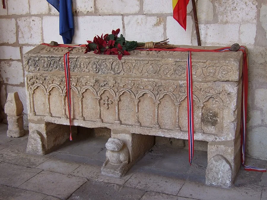 A carved stone sarcophagus tomb, arcaded along its sides, wrapped in Norwegian and Spanish ribbons with a bouquet of dark red flowers on top, standing against a stone cloister wall at the Colegiata de Covarrubias.