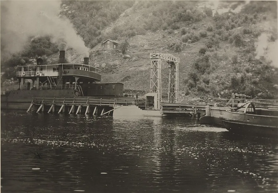 Black-and-white photograph of a steam railway ferry docked at a lakeside pier with a rail-loading gantry, smoke rising from its funnel, steep mountainous shoreline behind.