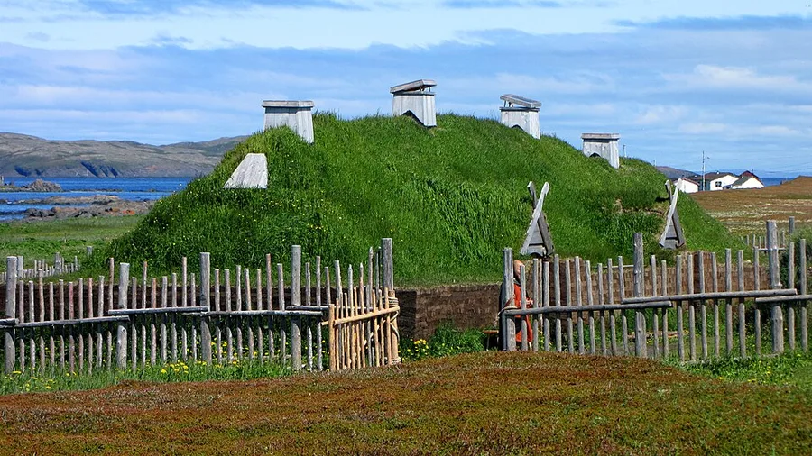 A grass-covered reconstructed Norse sod longhouse with a rounded turf roof and peaked wooden chimney vents, enclosed by a low post-and-wattle fence, on a coastal Newfoundland site with hills and water in the background.