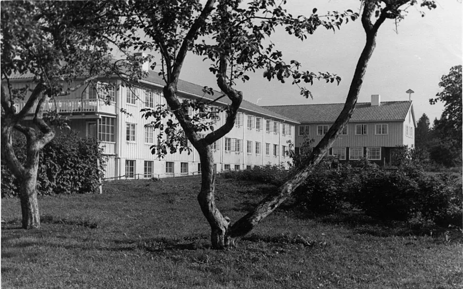 Black-and-white exterior photograph of a large multi-story wooden institutional building with balconies and many windows, seen across a lawn with fruit trees in the foreground.