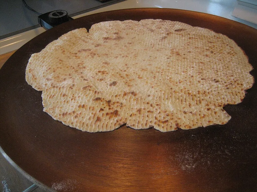 A round sheet of pale potato lefse, speckled with brown scorch marks, cooking on a large round electric griddle dusted with flour.