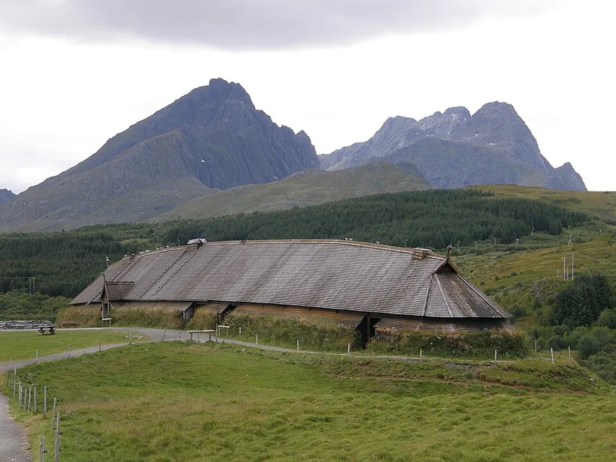 A long, grass-and-shingle-roofed reconstructed Viking longhouse stretches across a green hillside at Borg, Lofoten, with a cluster of jagged gray mountain peaks rising behind it under an overcast sky.