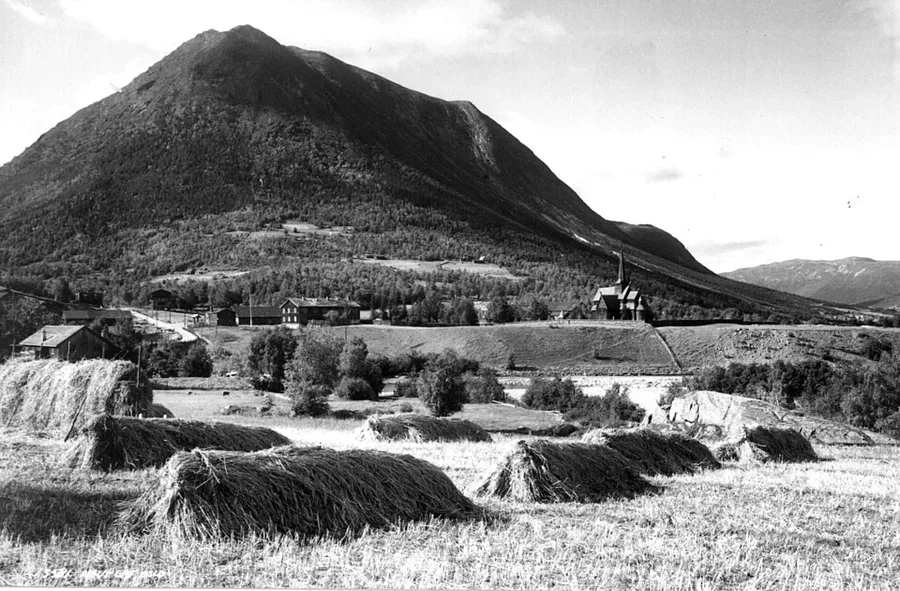 A 1935 black-and-white photograph of Lom Stave Church's tiered, dragon-finialed roof rising past a steep mountainside, with haystacks and mown hayfields in the foreground and farm buildings clustered near the church.