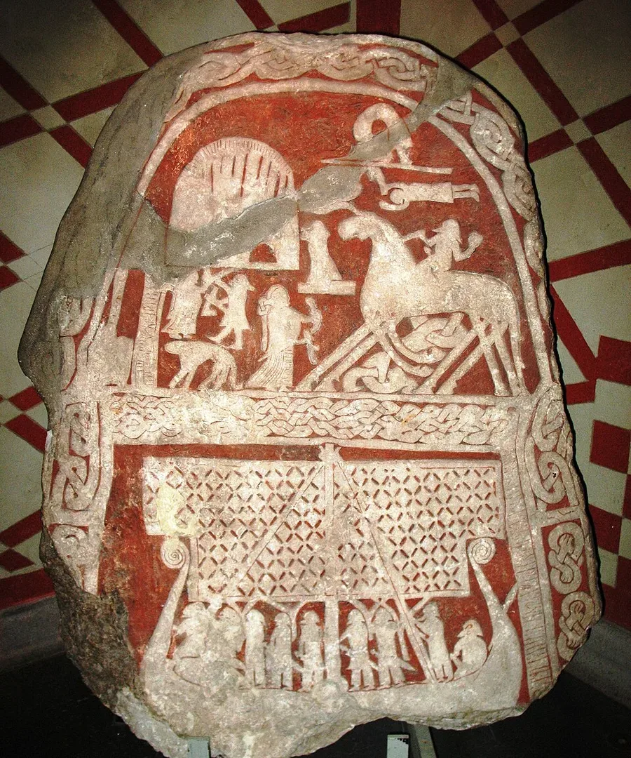 The Tjängvide image stone, Gotland (8th–9th c.): pale relief on red-painted stone — a rider on Odin’s eight-legged horse Sleipnir met by a figure bearing a drinking horn above, and below, a Viking longship under a broad diamond-patterned sail, crew ranged along the hull.