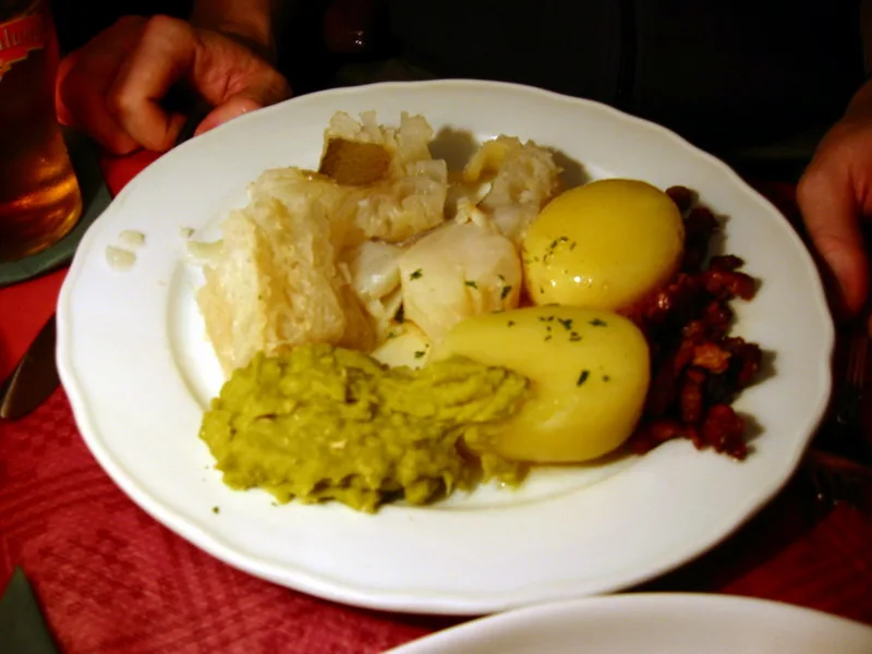A white plate of pale, gelatinous lutefisk pieces alongside boiled potatoes, mashed green peas, and crisp bacon-fat cracklings, set on a red tablecloth, a diner's hands and a glass of beer beyond.