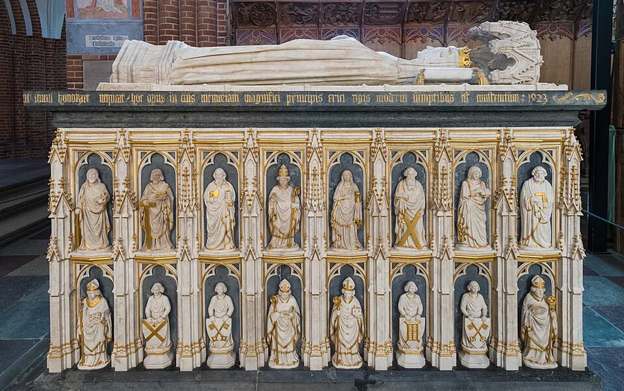 Gilt alabaster tomb chest and effigy of Queen Margrete I inside Roskilde Cathedral, the crowned recumbent figure on top and two tiers of gilded saint and bishop statuettes in arched niches on the sarcophagus front.
