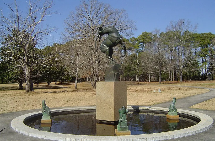 A bronze sculpture of a gazelle arched in mid-leap atop a stone pedestal at the center of a circular fountain, with three small bronze animal figures ringing the pool's edge, set in a grassy park with bare winter trees.