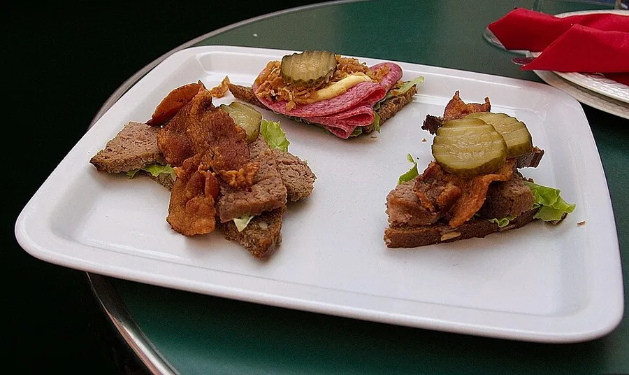 Three garnished open-faced slices of coarse dark bread on a white serving tray on a café table with a red napkin — pâté with crisp bacon, pickle, and lettuce on two slices, folded salami ruffles with crispy onion on the third.