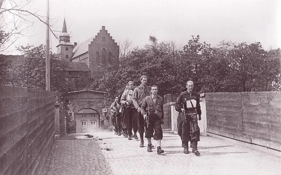Black-and-white 1945 photograph of a column of armed men in mixed civilian and military dress marching through a fortress gate at Akershus, a church tower rising behind the walls.