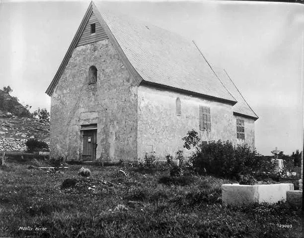 1926 black-and-white photograph of Moster gamle kyrkje, a small medieval stone church with a steep shingled roof, seen from the churchyard corner among grass and low shrubs; a photographic plate number is stamped in the bottom corner.