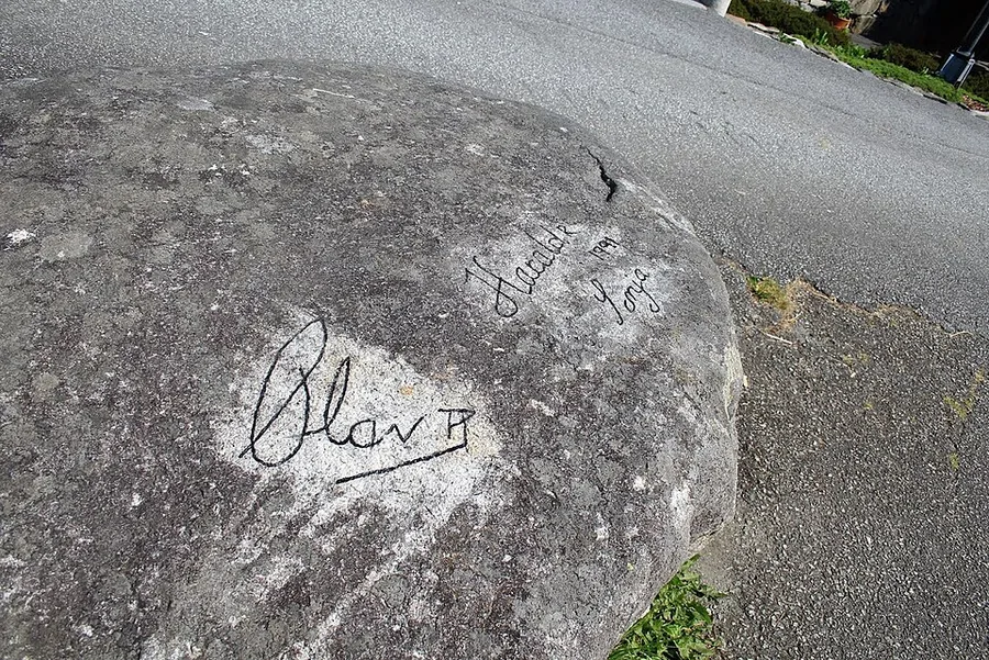 Kongesteinen, a smooth rounded boulder beside Moster old church, carved with royal signatures: "Olav" with a crown mark at lower left, and "Harald," "Sonja," and the date 1991 at upper right, marking royal visits to the assembly site.