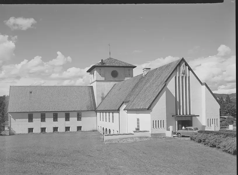 Black-and-white photograph of the Viking Ship House on Bygdøy: whitewashed gabled halls meet at a central square tower, the tall entrance facade striped with narrow vertical windows, a 1950s car parked by the door and open lawn in the foreground.