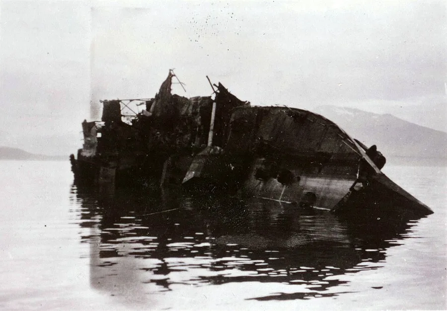 Black-and-white 1940 photograph of a wrecked German destroyer's broken hull listing in Narvik harbor, snow-capped mountains visible across the fjord.