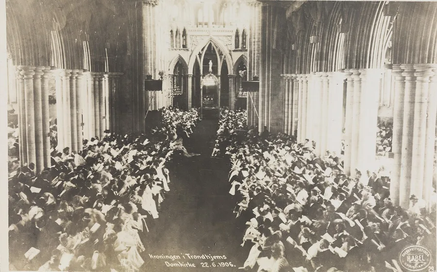 Sepia postcard photograph of the packed nave of Nidaros Cathedral during the coronation of Haakon VII: the congregation fills the pews between massive gothic piers, the choir screen beyond, with the caption 'Kroningen i Trondhjems Domkirke 22.6.1906' at the bottom.