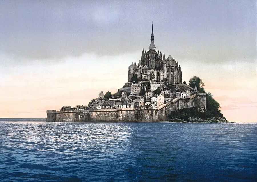 Mont-Saint-Michel at high water, c. 1900: the abbey and its slender Gothic spire crown the rock, terraced village houses and long rampart walls below, the whole mount ringed by blue sea under a pale sky — a hand-colored photochrom-era view.